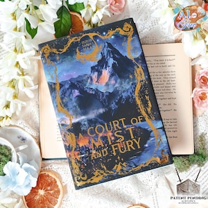 Könnte beinhalten: Ein Buch mit dem Titel "A Court of Mist and Fury" mit einem dunkelblauen Einband mit einer Berglandschaft und goldfarbenen Buchstaben. Das Buch ist auf einem alten Buch mit Text geöffnet. Dekorative Blumen und eine Teetasse umgeben das Buch.