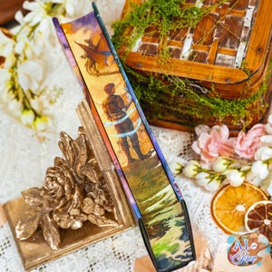 May include: A gold floral bookend supporting a book with a fantasy illustration on its spine. The book's spine shows a figure, a dragon, and a landscape. The background includes a wooden box and floral decorations. The bookend is made of metal.