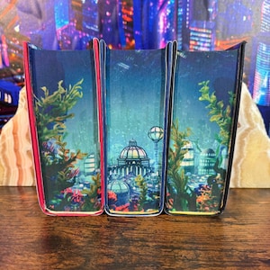 May include: Three decorative bookends with a blue, green, and red background. Each bookend features a cityscape with a dome-shaped building and a tall tower. The bookends are made of a hard material and have a glossy finish.