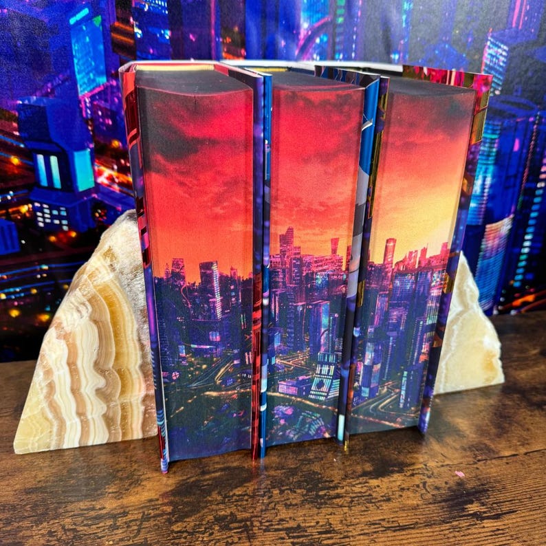May include: Three bookends with a cityscape design. The bookends are made of a hard material and have a glossy finish. The cityscape is a mix of red, orange, blue, and purple colors. The bookends are on a wooden surface.