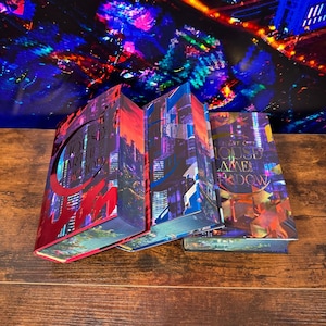 May include: Three hardcover books with colorful cityscapes on the covers. The titles are "House of Earth and Blood", "House of Sky and Breath", and "House of Chains and Shadow".