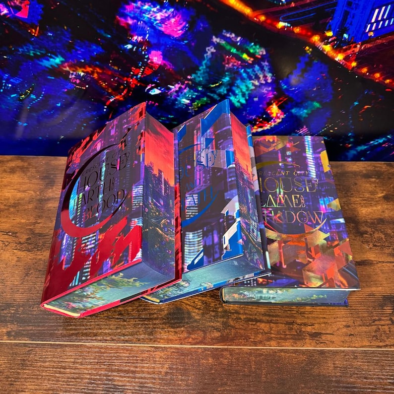 May include: Three hardcover books with colorful cityscapes on the covers. The titles are "House of Earth and Blood", "House of Sky and Breath", and "House of Chains and Shadow".