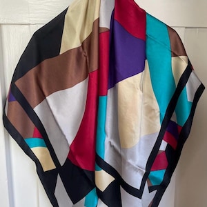 Peut inclure: Un foulard carré présentant un motif géométrique audacieux avec des blocs de couleurs incluant le rouge profond, le turquoise vif, le violet, le noir, la crème et le marron. Le design se compose de formes et de lignes angulaires nettes.