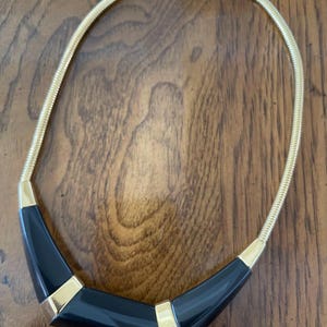 May include: A gold-tone snake chain necklace featuring a central curved pendant composed of three smooth black resin segments separated by two polished gold-tone metal accents. The necklace includes a lobster claw clasp and a small dangling charm.