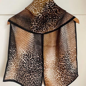 May include: A silk scarf with a leopard print design in shades of brown, black, and tan. The scarf has a black border and is displayed on a wooden hanger.