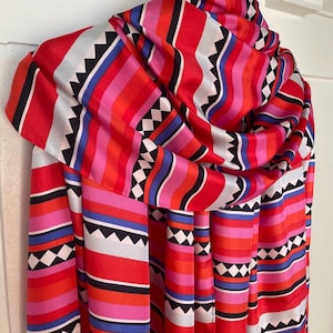 May include: A rectangular scarf featuring a vibrant pattern of horizontal stripes in shades of red, pink, blue, white, and black. The design includes repeating geometric bands with black sawtooth edges and white diamond shapes on a black background.
