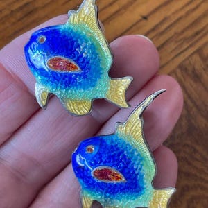 May include: A pair of fish-shaped earrings. Each earring has a blue body with yellow and green accents, a red oval detail, and a silver outline. The earrings are held in a person's hand.