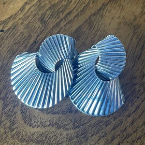 May include: A pair of silver earrings. Each earring has a fan-shaped design with vertical lines radiating from a central opening. The earrings are presented on a wooden surface.