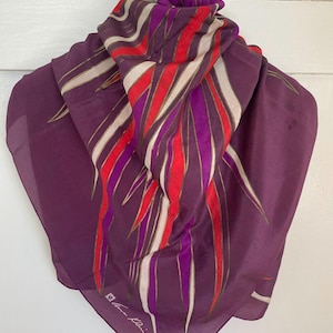May include: A square scarf featuring a deep purple base with a bold, abstract pattern of elongated, pointed shapes in vibrant red, bright magenta, and cream. The design radiates from the centre, and the corner displays the signature Anne Klein logo.