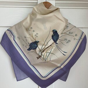 May include: A square silk scarf with a cream background and a lavender border. The scarf features a design of two blue birds perched on a branch with green foliage. The scarf is displayed on a wooden hanger.