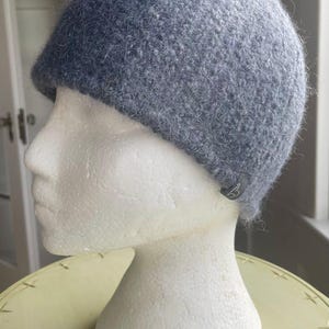 May include: A solid grey beanie with a fuzzy, textured knit surface. The hat features a rounded crown and a small, dark grey fabric tag attached to the lower edge, which displays a white stylized letter logo on its visible surface.