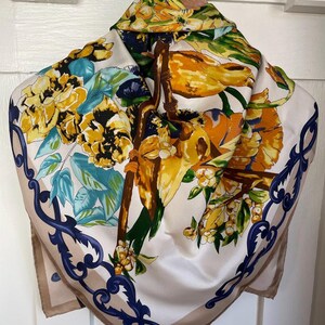 May include: A square scarf featuring a cream background with a vibrant print of yellow flowers, green leaves, and brown branches. The border is decorated with a dark blue ornate scrollwork pattern, framing the central botanical illustration.