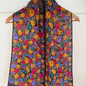 May include: A silk scarf with a dark blue background and a pattern of colourful gemstones. The gemstones are in shades of red, yellow, green, and purple. The scarf is fringed on the edges.