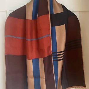 May include: A rectangular scarf featuring a geometric pattern of bold blocks and stripes in shades of rust orange, deep brown, tan, and royal blue. The design includes thin black horizontal lines on the right side and a light tan hem with a fringed edge.