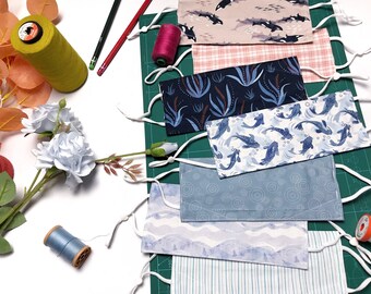May include: A collection of six fabric face masks with different patterns, including fish, plants, and geometric designs. The masks are laid out on a green cutting mat with white thread and spools of thread.