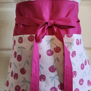 May include: A pink and white apron with a cherry pattern. The apron has a wide pink waistband that ties in the back. The cherry pattern features pink cherries with green stems and leaves. The apron is displayed on a black metal stand.