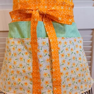 May include: A green and orange apron with a floral print and a tie. The apron has two pockets and a wide waistband.