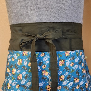 May include: A waist apron with a dark green waistband and ties. The lower portion features a blue floral pattern with elephants and flowers. The apron is tied in a bow at the front.