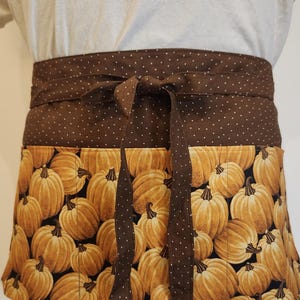 May include: A waist apron featuring a brown fabric waistband with a white polka dot pattern and a tie closure. The lower section of the apron displays a repeating print of orange pumpkins on a black background, complete with visible vertical stitching.