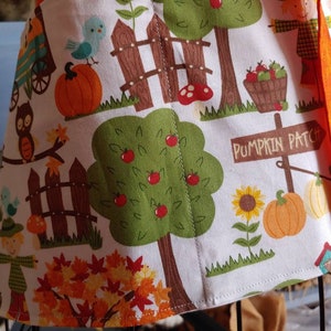 Op de afbeelding: Een wit schort met een kleurrijke herfstprint van pompoenen, appelbomen, vogelverschrikkers, zonnebloemen en een houten bord met de tekst PUMPKIN PATCH. Het schort is voorzien van feloranje stoffen linten en een oogsttafereel.