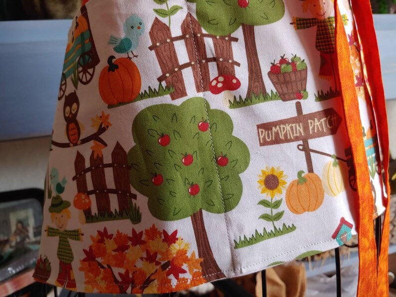 Op de afbeelding: Een wit schort met een kleurrijke herfstprint van pompoenen, appelbomen, vogelverschrikkers, zonnebloemen en een houten bord met de tekst PUMPKIN PATCH. Het schort is voorzien van feloranje stoffen linten en een oogsttafereel.
