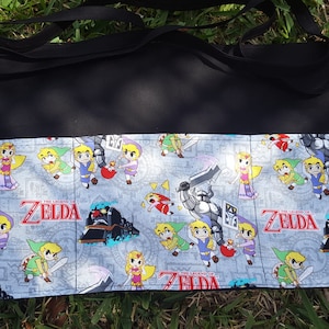 May include: A black apron with a multi-colored fabric pocket featuring a repeating pattern of cartoon characters from the Legend of Zelda video game series. The fabric pocket has the words "The Legend of Zelda" printed on it.