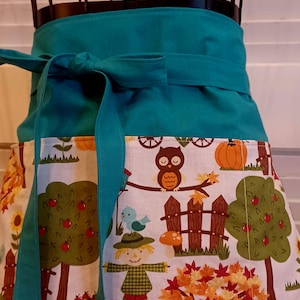 Op de afbeelding: Een blauwgroen tailleschort met een wit stoffen paneel voorzien van een kleurrijke herfstprint. Het patroon toont uilen, vogelverschrikkers, pompoenen, appelbomen en een bordje met de tekst PUMPKIN PATCH. Het schort heeft een effen blauwgroene tailleband.