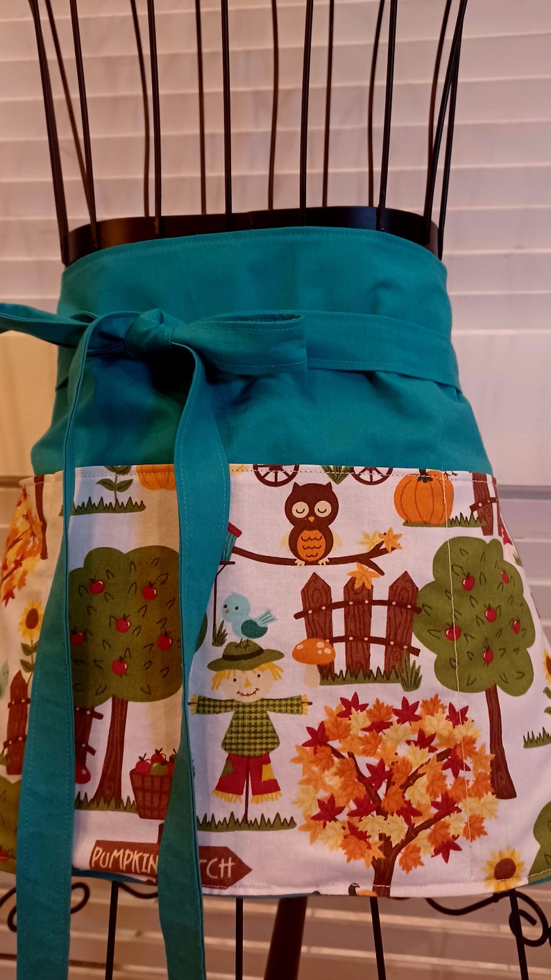 Op de afbeelding: Een blauwgroen tailleschort met een wit stoffen paneel voorzien van een kleurrijke herfstprint. Het patroon toont uilen, vogelverschrikkers, pompoenen, appelbomen en een bordje met de tekst PUMPKIN PATCH. Het schort heeft een effen blauwgroene tailleband.