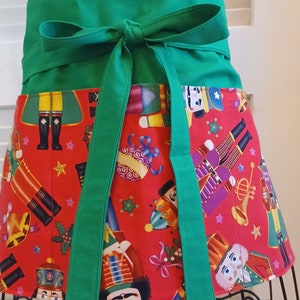 May include: A festive half apron with a green waistband and ties. The apron's lower portion features a red background adorned with colorful nutcracker figures, stars, and holiday-themed illustrations. The apron is displayed on a black metal stand.