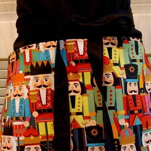 May include: A black apron with a colorful pattern of nutcracker soldiers. The apron features a black waistband and ties. The nutcracker design includes red, yellow, green, and blue uniforms with various facial expressions.