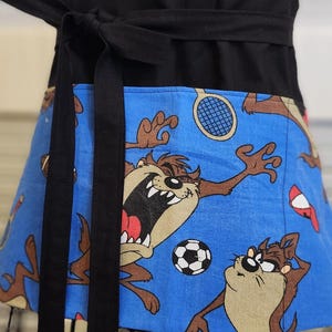 May include: A blue and black apron with a cartoon character design. The apron has a black waistband and a large pocket on the front. The cartoon character is Taz from Looney Tunes, and he is playing sports.