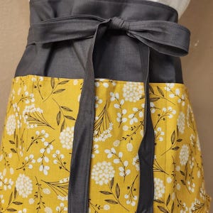 May include: A yellow apron with a white floral pattern and gray ties. The apron has a gray waistband and a pocket on the front.
