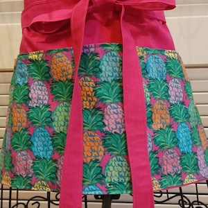 May include: A pink apron with a pineapple print and a matching pink tie. The apron has a large pocket on the front.