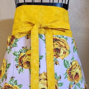 May include: A yellow floral print apron with a yellow waist tie. The apron has a white background with yellow roses and green leaves.