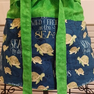 May include: Green and blue fabric apron with a tie waist and a pocket. The apron has a sea turtle design and the text "Wild & Free as the Sea".