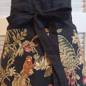 May include: Black apron with a jungle print featuring monkeys and tropical leaves. The apron has a black tie that can be tied in the front.