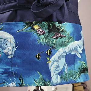 May include: A navy blue waist apron featuring a large front pocket with a vibrant underwater print. The fabric displays light gray manatees, yellow and black angelfish, green aquatic plants, and seashells against a deep blue water background.