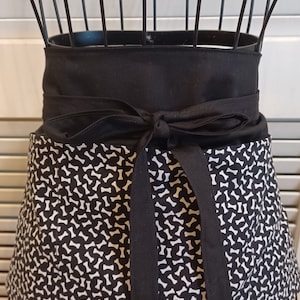 May include: Black and white dog bone print apron with a solid black waistband and long ties. The apron is displayed on a wire dress form. The apron's design features a repeating pattern of white dog bones on a black background.