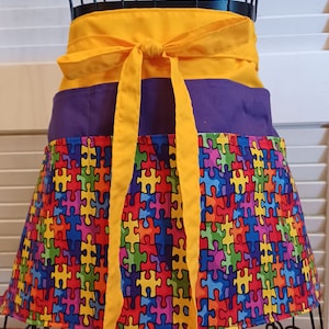 May include: A yellow and purple apron with a colorful puzzle piece pattern. The apron has a yellow tie and a purple waistband.