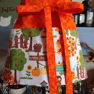 May include: A half apron featuring a solid orange waistband with long ties and a white skirt section printed with a colorful autumn scene. The pattern includes pumpkins, sunflowers, trees, a wooden fence, and a sign that reads PUMPKIN PATCH.