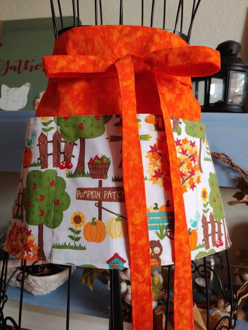 Op de afbeelding: Een halfschort met een effen oranje tailleband met lange linten en een wit rokgedeelte bedrukt met een kleurrijke herfstsc&egrave;ne. Het patroon bevat pompoenen, zonnebloemen, bomen, een houten hek en een bord met de tekst PUMPKIN PATCH.