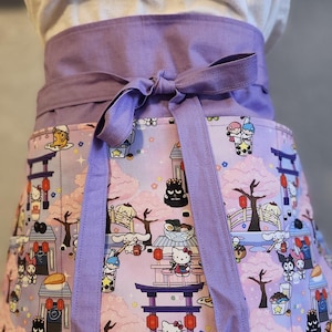 May include: A purple apron with adjustable ties. The apron has a pink and white print featuring Hello Kitty and other characters in a Japanese-inspired setting. The print includes cherry blossoms, lanterns, and a temple.
