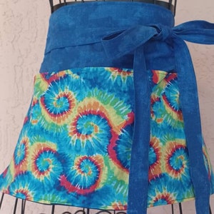 May include: A blue tie-dye apron with a matching blue waist tie. The apron features a colorful spiral pattern in shades of blue, yellow, orange, and red. The apron is displayed on a black metal form.