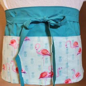 May include: A teal waist apron with a flamingo and pineapple print. The apron has a teal waistband and ties, and a pocket with a light blue background. The flamingos are pink and the pineapples are light blue.