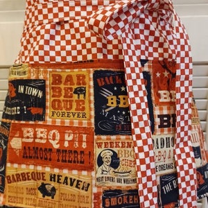 May include: A red and white checkered apron with a pocket featuring a variety of BBQ themed designs. The designs include text such as "Barbeque Heaven", "BBQ Pit Almost There", and "Backyard BBQ".