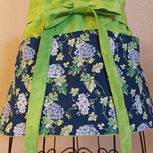 May include: A waist apron with a vibrant floral pattern. The apron features a navy blue background adorned with light purple and white hydrangea blooms and green leaves. The apron has a bright green waistband and long ties.