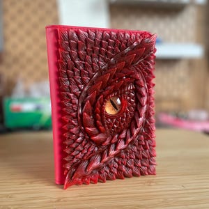Peut inclure: Un journal rouge sur le thème de la fantaisie avec un motif d'œil de dragon. La couverture présente un motif texturé et détaillé d'écailles rouges et un œil de dragon central avec des détails orange et noirs. Le journal est posé sur une surface en bois.
