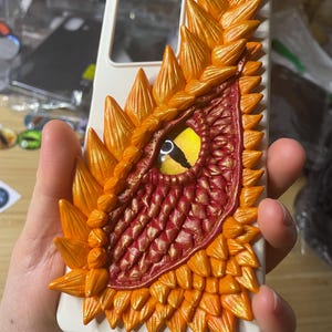 May include: A white phone case featuring a detailed, three-dimensional dragon eye design. The eye is yellow with a black slit pupil, surrounded by red and gold scales and orange, spiky accents. The case is held in a hand.