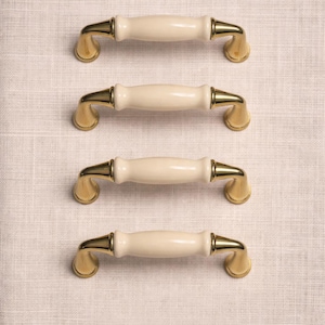 May include: Four cabinet pulls with a cream-colored, cylindrical center and gold-toned metal accents. The pulls are arranged in a vertical row against a neutral, textured background. These are likely for furniture or cabinetry.