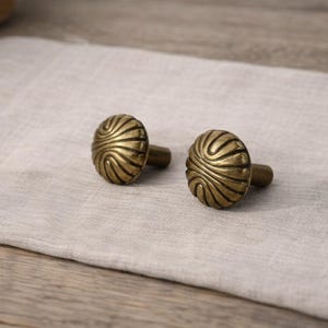 Könnte beinhalten: Zwei antike Messing-Schrankknöpfe mit einem Wirbeldesign. Die ovalen Knöpfe haben einen warmen, goldenen Farbton. Sie liegen auf einem neutralen Leinentuch, was auf eine Vintage- oder rustikale Ästhetik hindeutet.